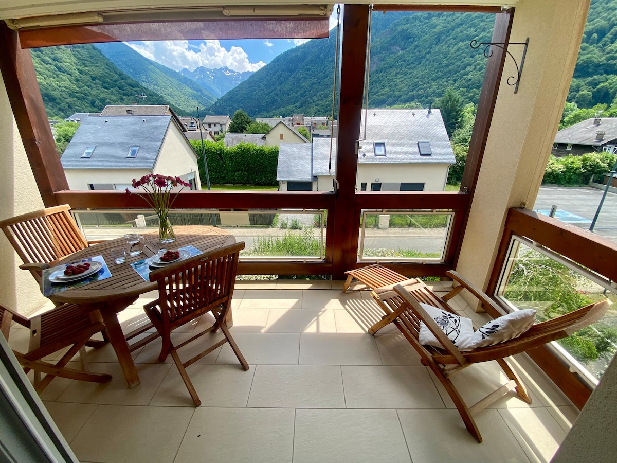 A covered balcony features a wooden table set for two, with a vase of flowers in the center. Two lounge chairs are positioned to enjoy expansive mountain views through large windows that fill the space with natural light.