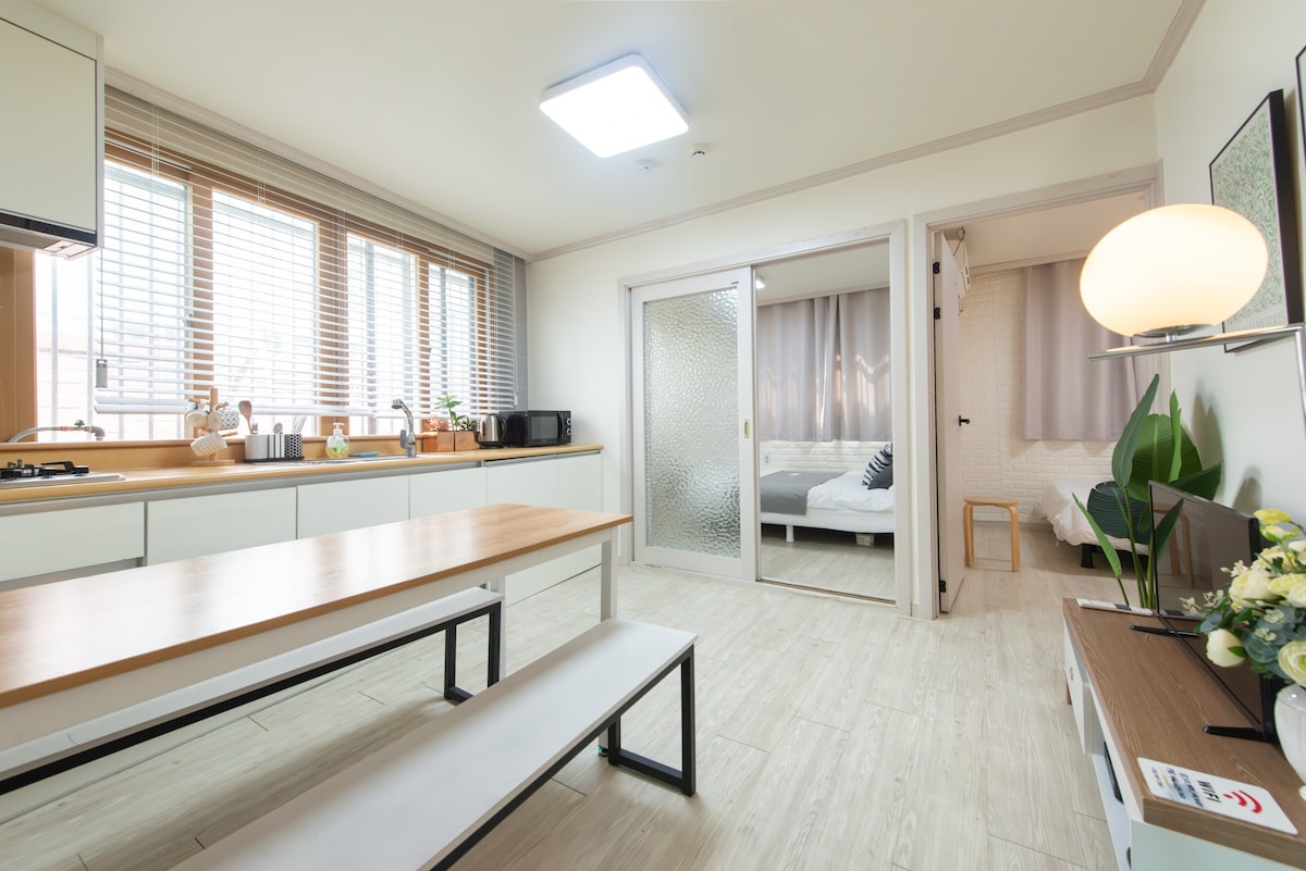 A modern kitchen area is visible, featuring light-colored cabinetry and a wooden table with benches. Large windows provide natural light, and nearby appliances include a microwave and a kettle. A sliding door can be seen leading to a bedroom, partially revealing the cozy sleeping space.