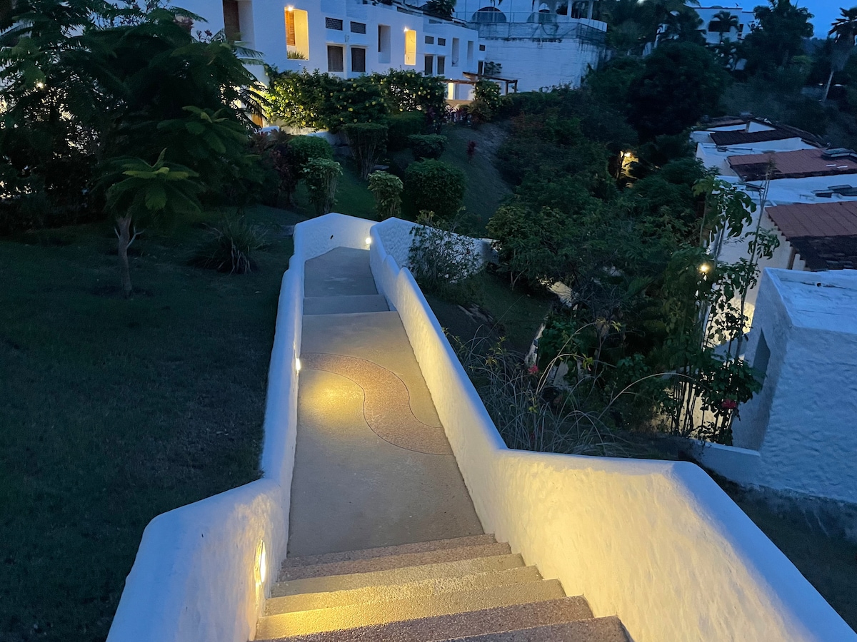 A well-lit pathway descends through lush greenery, leading to whitewashed buildings. Soft lighting along the steps highlights the curved design of the pathway, creating a sense of tranquility in the evening atmosphere.