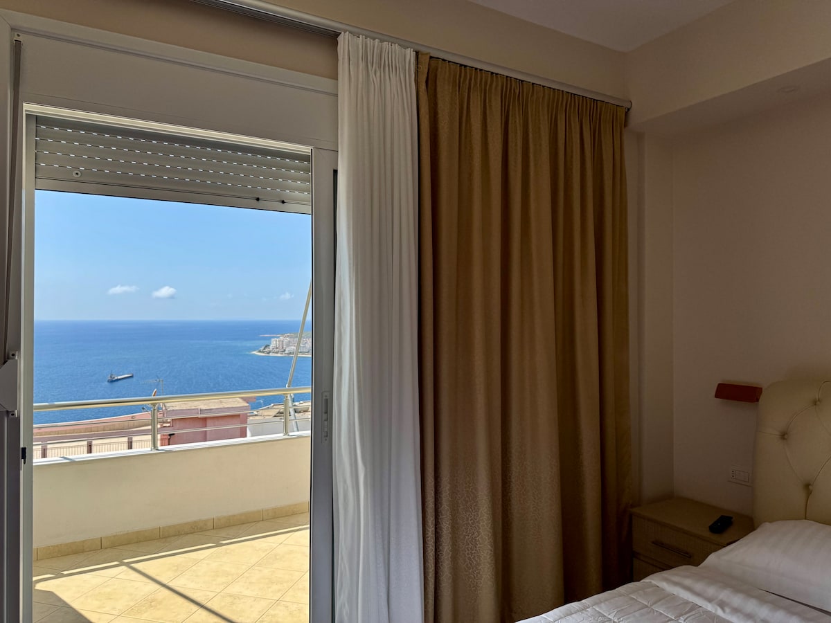A spacious room is highlighted by a large window and glass door that open to a balcony. The balcony provides expansive views of the blue sea. Soft curtains frame the entrance, allowing natural light to enter the interior.