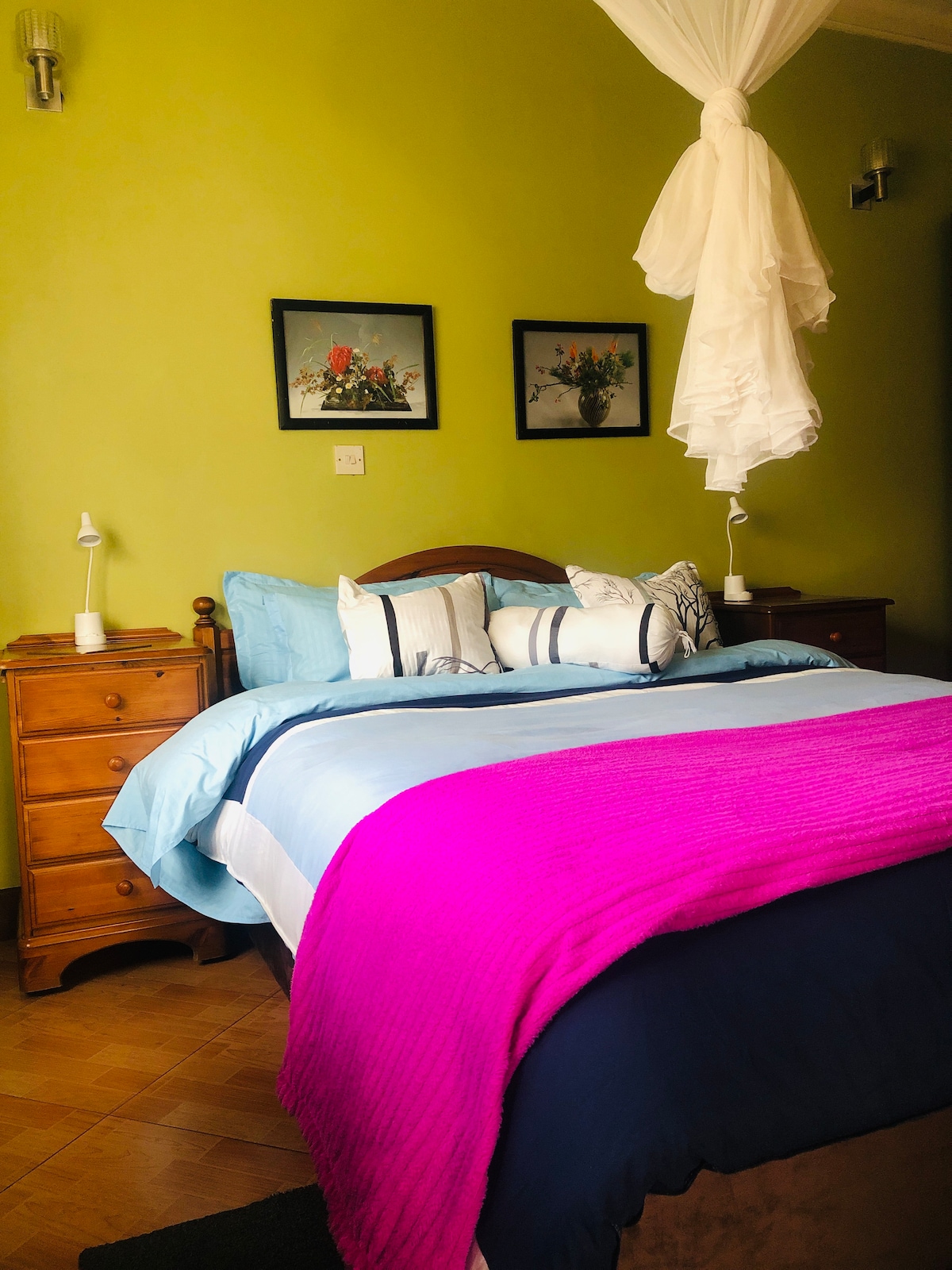 A cozy sleeping area features a spacious bed with layered bedding in shades of blue and purple. Two wooden nightstands with lamps sit beside the bed, and floral artwork adorns the green walls, enhancing the warm ambiance.