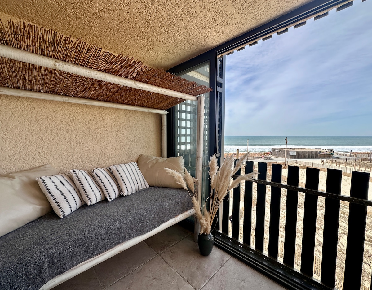 A cozy seating area on a balcony features a built-in daybed with plush cushions. Natural elements are incorporated with decorative plants and a view of the ocean and beach visible through large sliding glass doors.