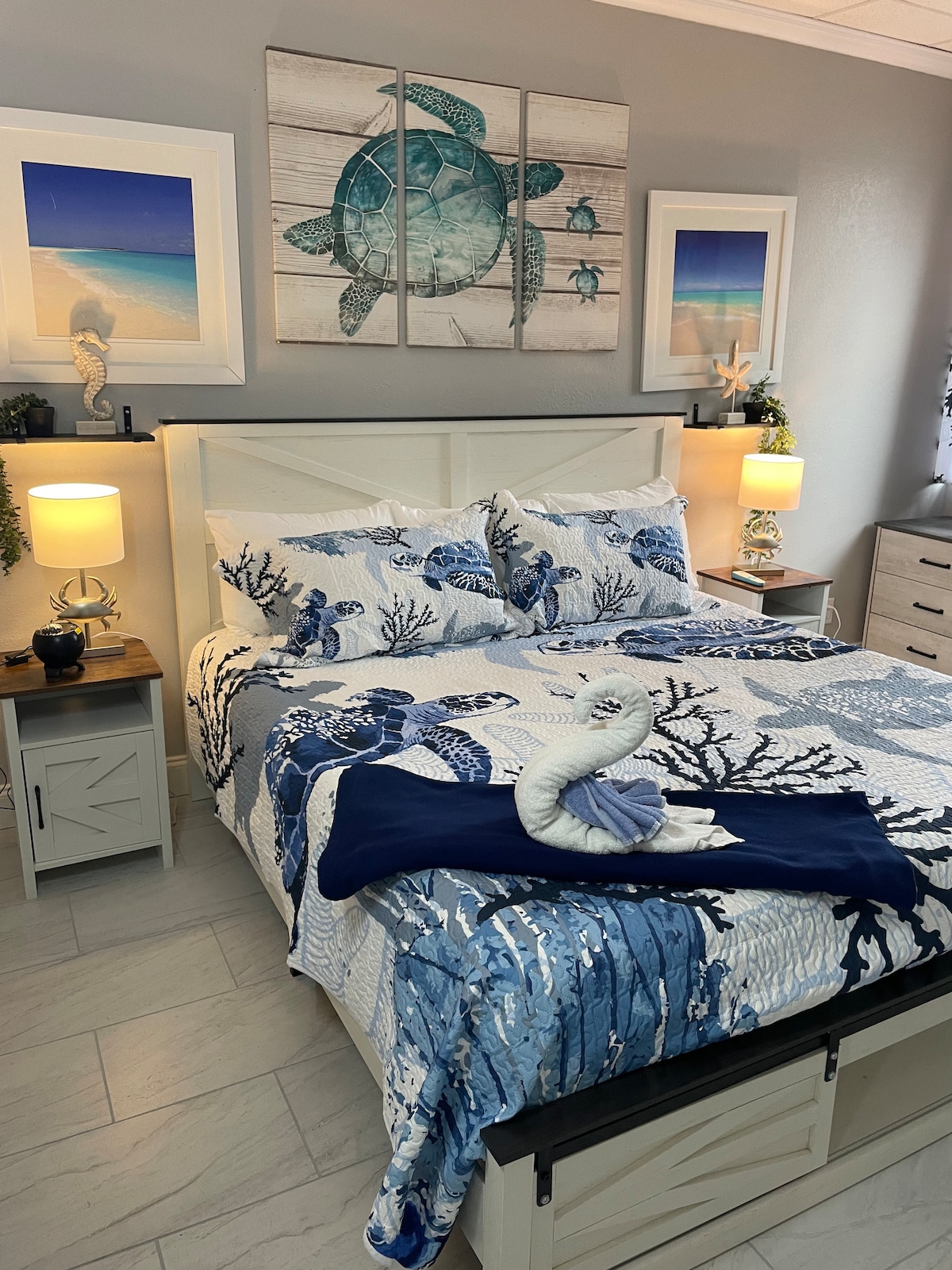 A serene sleeping area features a king bed with a coral-themed quilt and coordinating decorative pillows. Artistic sea turtle wall art adds coastal charm, complemented by two bedside lamps on matching nightstands. Natural light fills the space through a nearby window, enhancing the relaxing atmosphere.