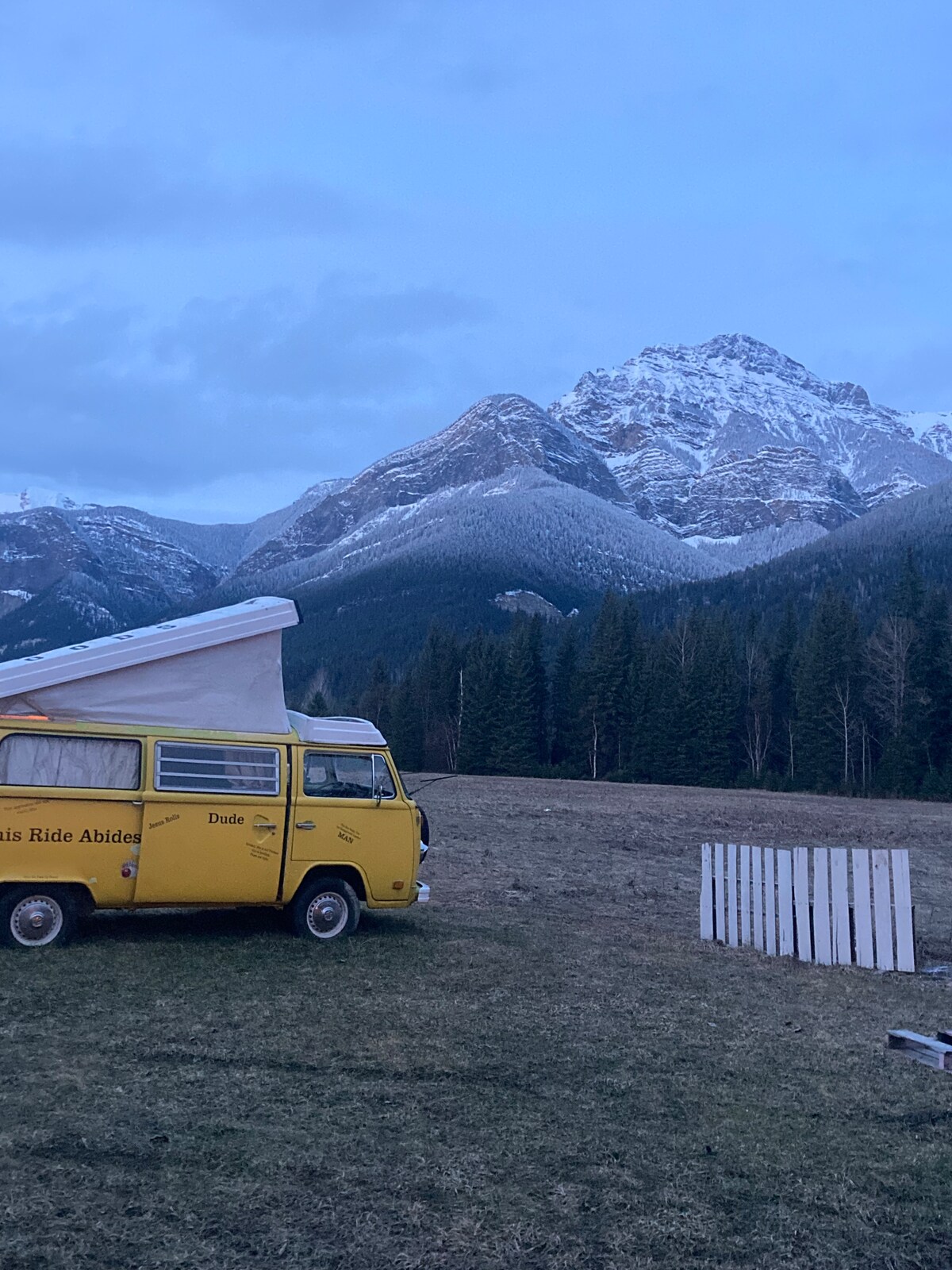 A vintage yellow bus is parked in an open field, with a backdrop of snow-capped mountains and a forest. The bus's pop-up top is extended, and a white wooden fence lines part of the scene. The sky displays a soft blue at dusk.