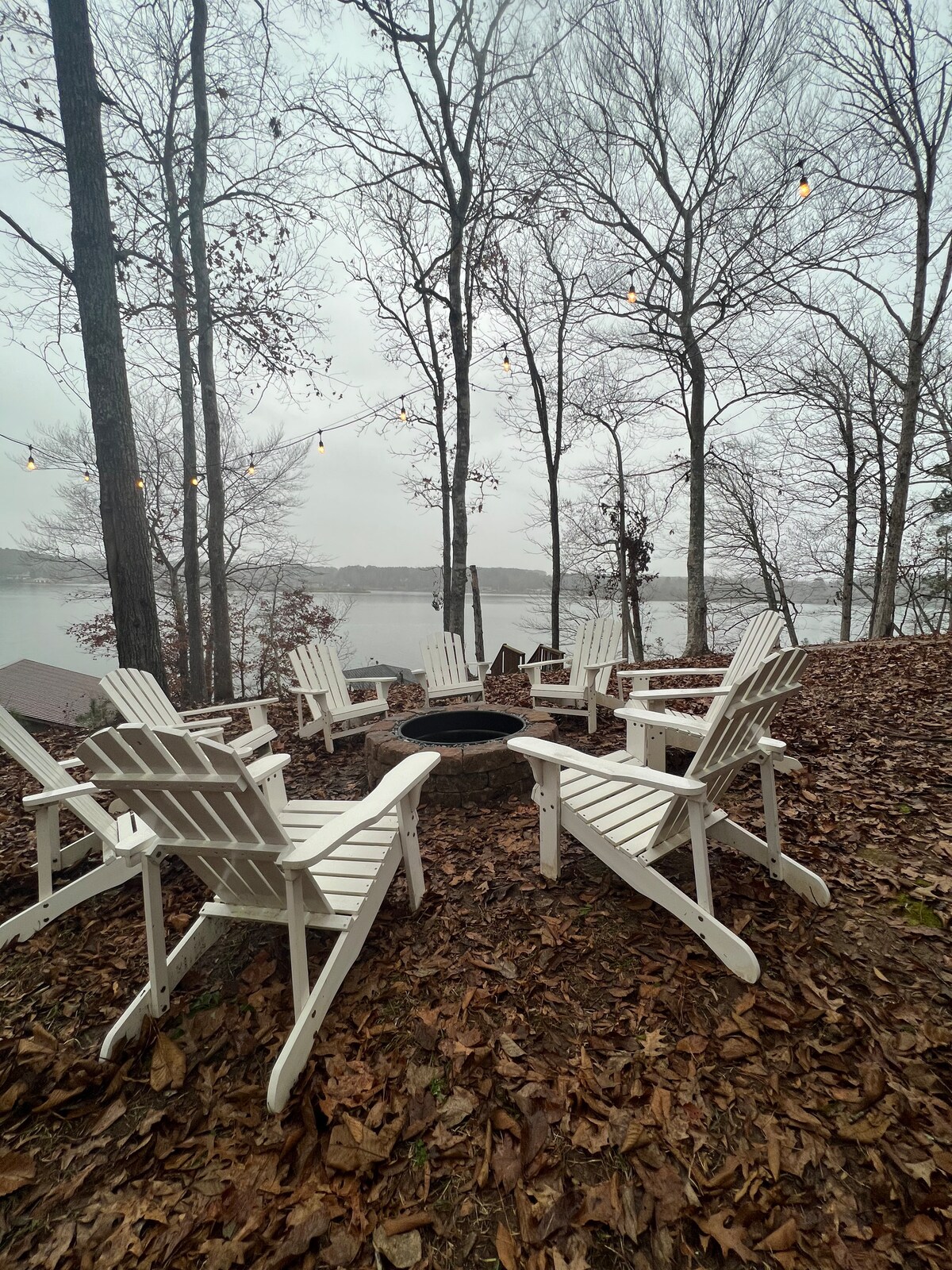 A circle of six white Adirondack chairs surrounds a fire pit, set among a bed of fallen leaves. Bare trees rise in the background, and the lake is visible in the distance, contributing to a serene, natural setting.