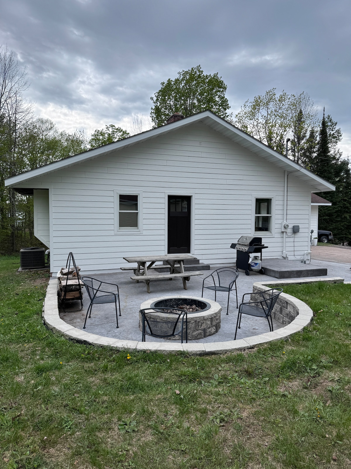 An outdoor patio area features a circular stone fire pit surrounded by six black chairs. A picnic table is positioned nearby, with a grill visible to the side. The home’s white exterior contrasts with the natural greenery of the yard.