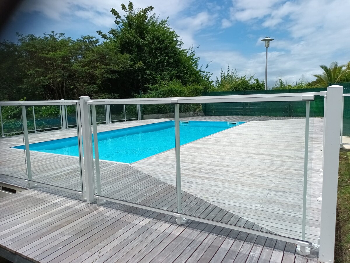 A swimming pool is surrounded by a wooden deck, featuring a transparent safety railing. Lush greenery is visible in the background, and the sky is partly cloudy, allowing natural light to brighten the pool's clear blue water.