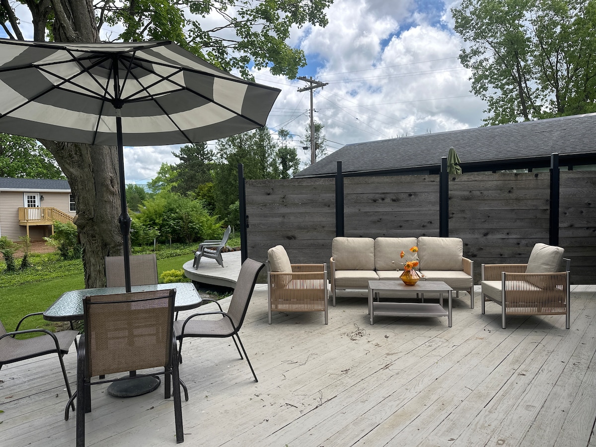A spacious deck features a seating area with a sectional sofa and decorative pillows, complemented by a small table with two chairs. An umbrella provides shade over the dining area, while a landscape of lush greenery is visible in the background.