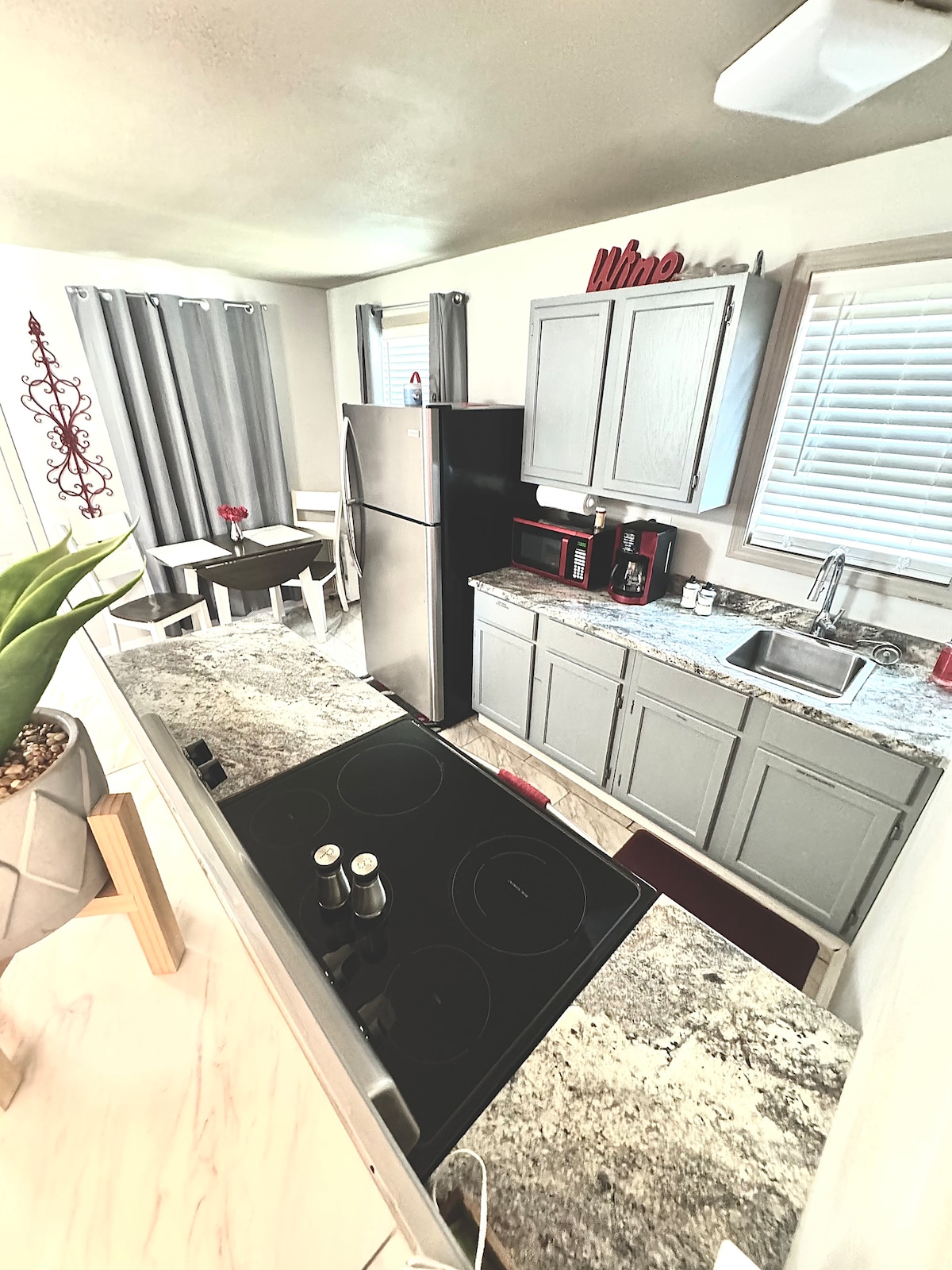 A functional kitchen is equipped with stainless steel appliances and gray cabinets. A small dining table with two chairs is positioned near a window with light curtains. The countertops are finished in a sleek gray stone, complementing the overall design.