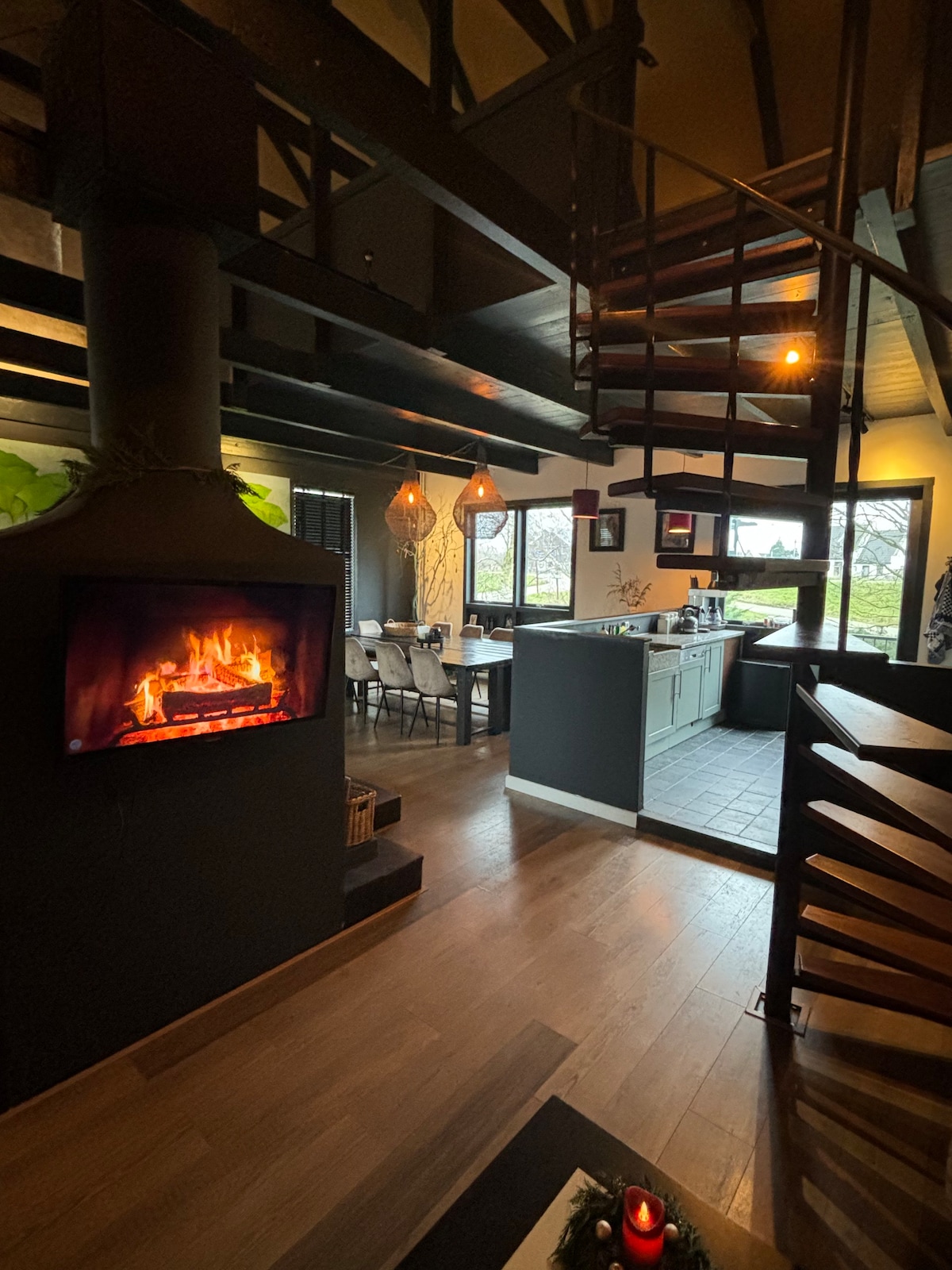 The interior space features a spiral staircase leading to an upper level, with a cozy fireplace mounted on the wall, casting a warm glow. An open kitchen is visible, equipped with modern appliances, while a dining area with multiple chairs is arranged near large windows offering views of the outdoors.