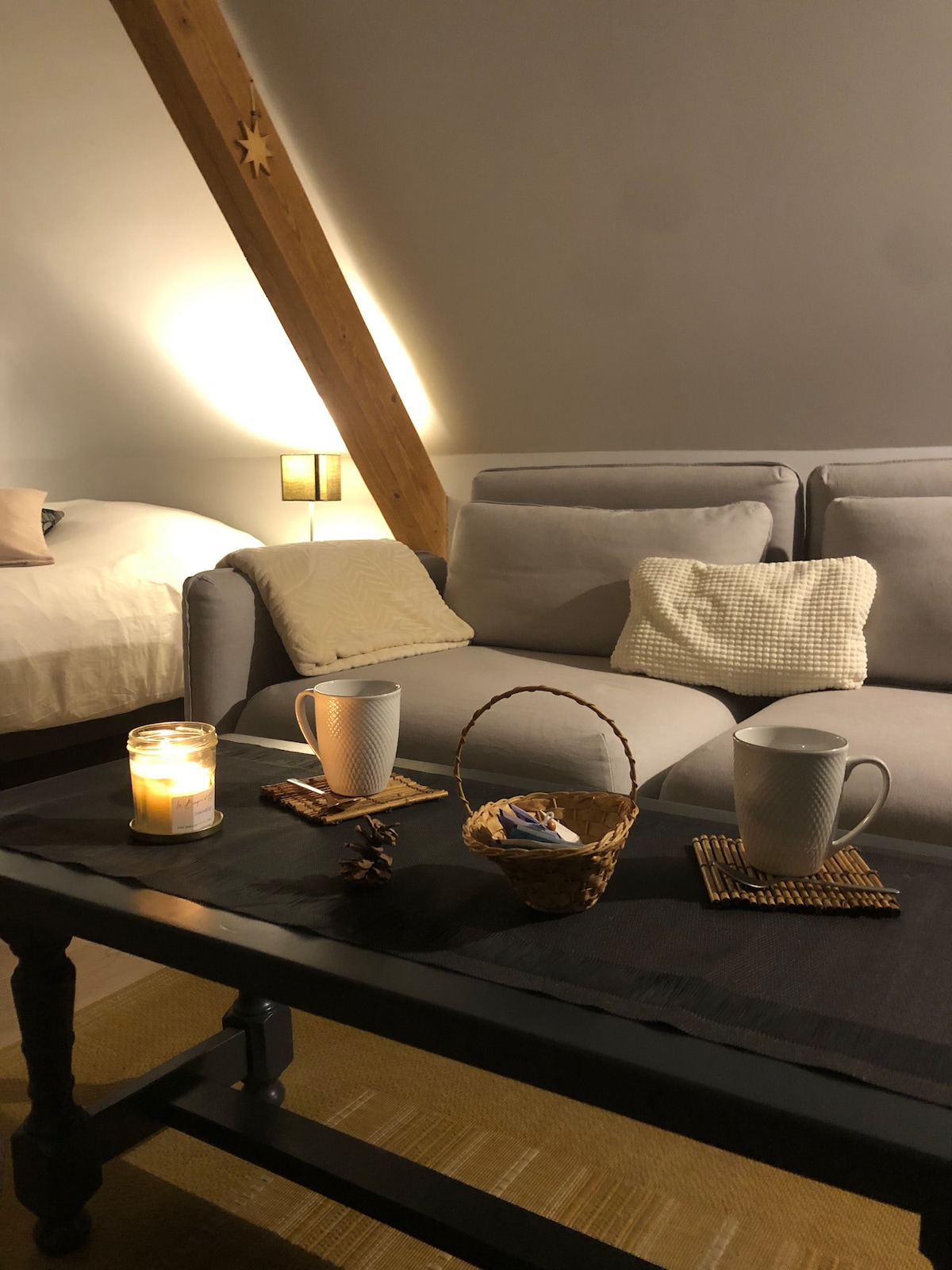 A cozy living area features a light grey sofa adorned with decorative cushions. A wooden table is set with two mugs and a woven basket, enhancing the warm atmosphere. Soft lighting highlights the exposed beams and creates an inviting ambiance.