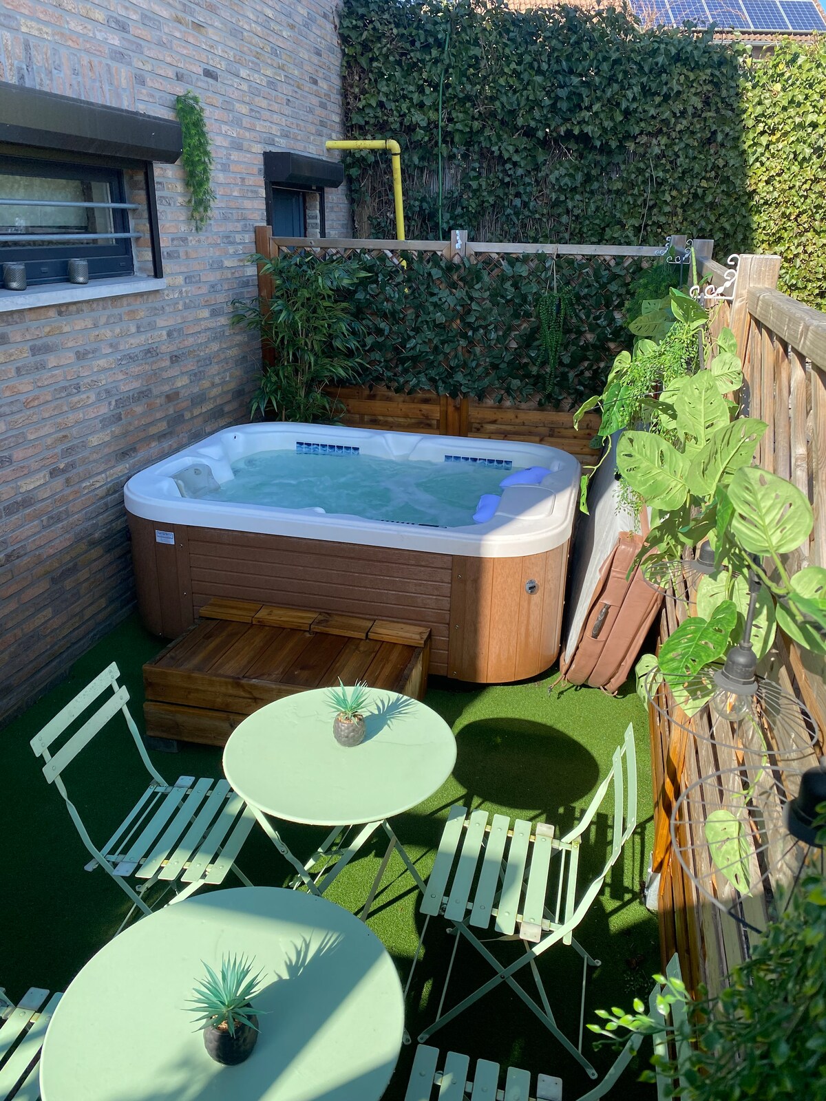 A private outdoor area features a hot tub surrounded by wooden privacy fencing and greenery. Two small green tables with matching chairs are positioned on a grass-like surface, creating a relaxed setting for outdoor enjoyment.