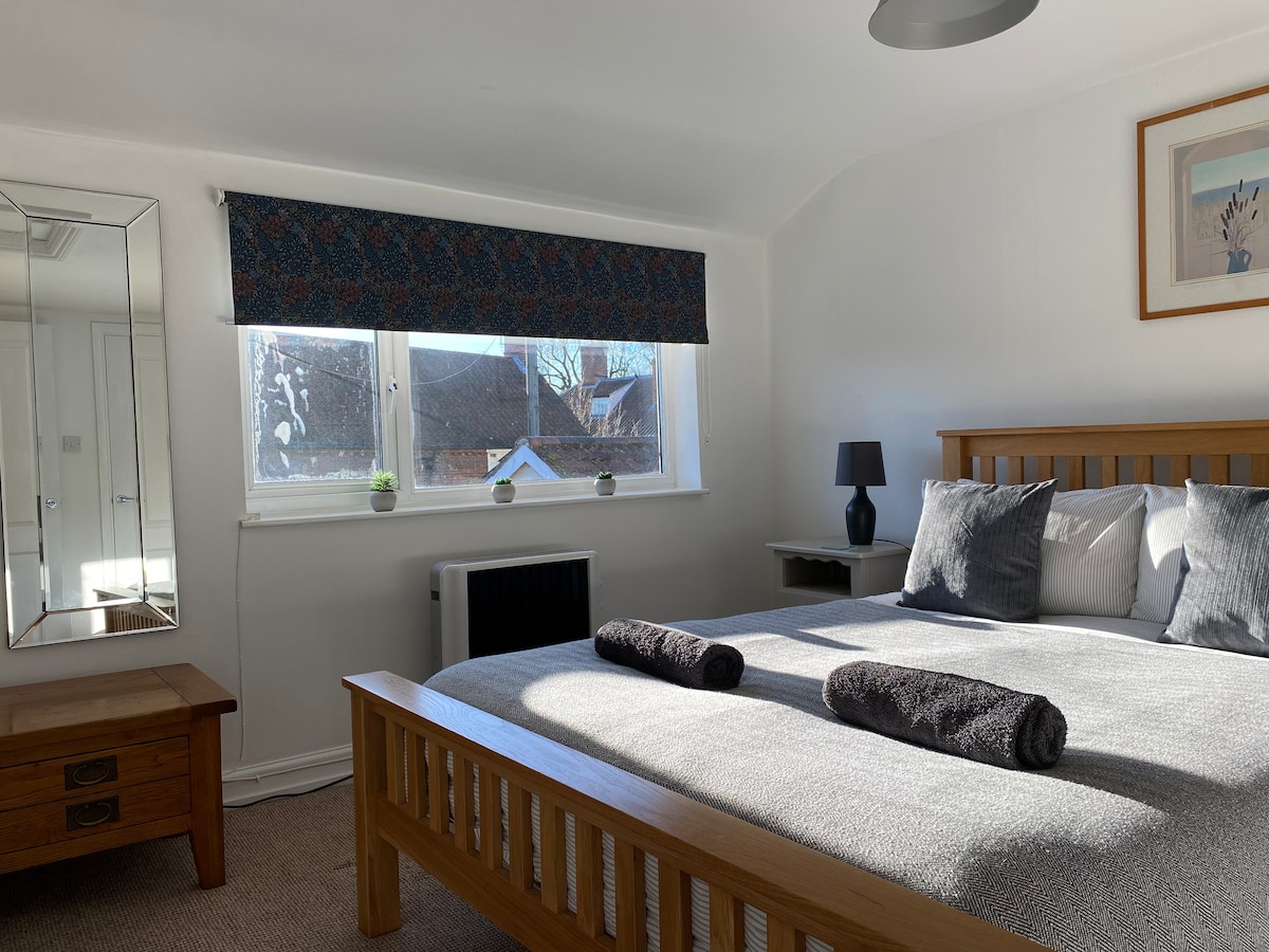 The spacious bedroom features a king-size bed adorned with soft pillows and towels. A large window allows natural light to brighten the room, while a mirror reflects the inviting space. A bedside lamp and a simple piece of wall art complete the minimalist decor.