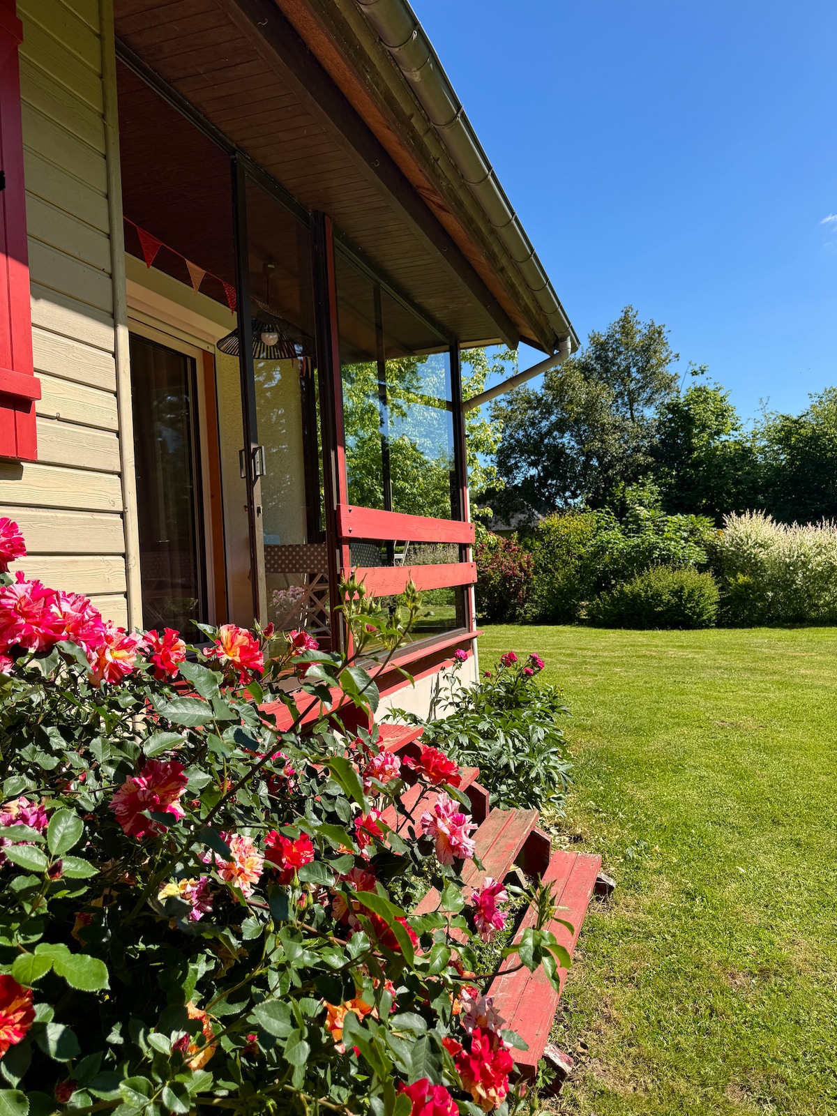 The exterior of a charming house is framed by colorful rose bushes and well-maintained grass. The entrance features a set of steps leading to a porch with glass panels, allowing for a view of the surrounding greenery under a clear blue sky.