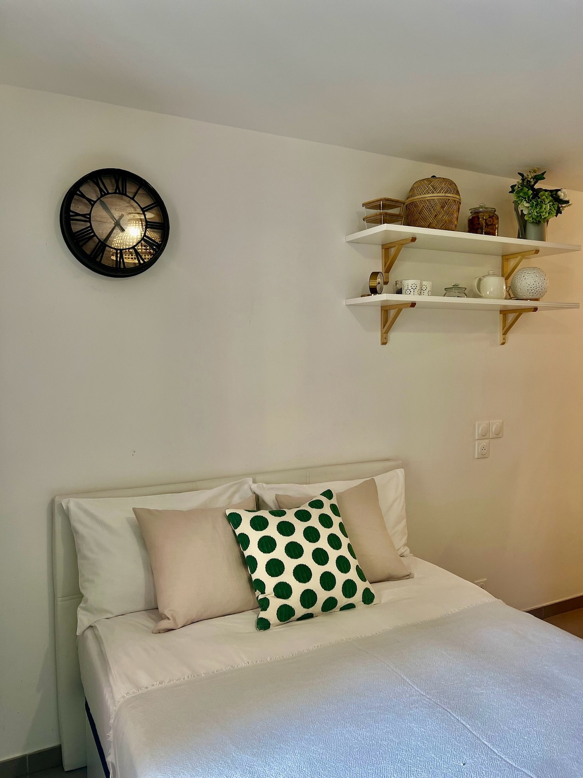 A simple bedroom is presented, featuring a spacious bed with soft white bedding. A decorative pillow with green polka dots rests atop the bed. Shelves above the bed display various decorative items, and a round wall clock is mounted nearby.