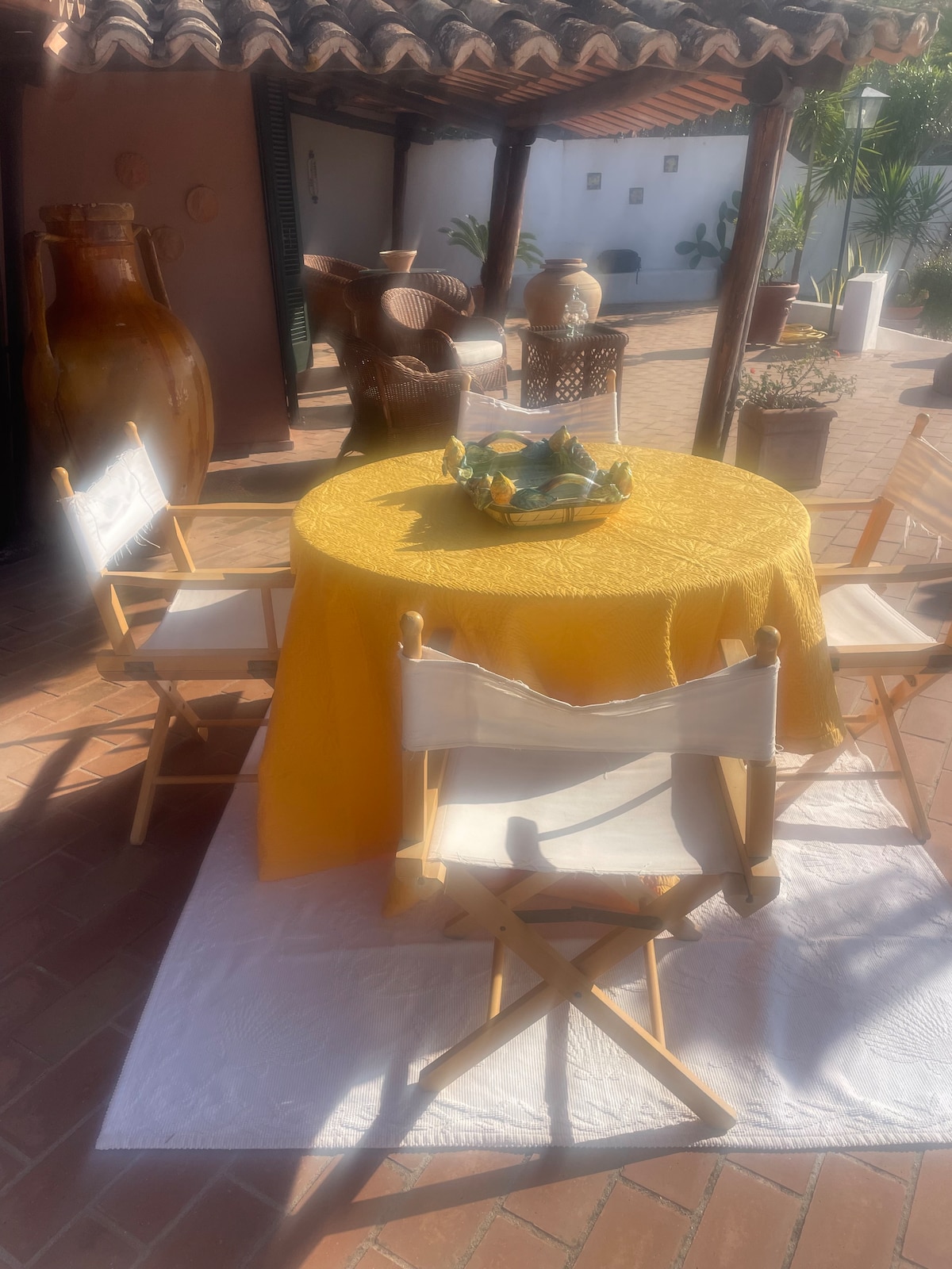 An outdoor dining area is highlighted with a round table covered in a yellow tablecloth. Four white, folding director's chairs surround the table. Decorative pottery and sculptures are arranged in the background, contributing to a vibrant, artistic setting.
