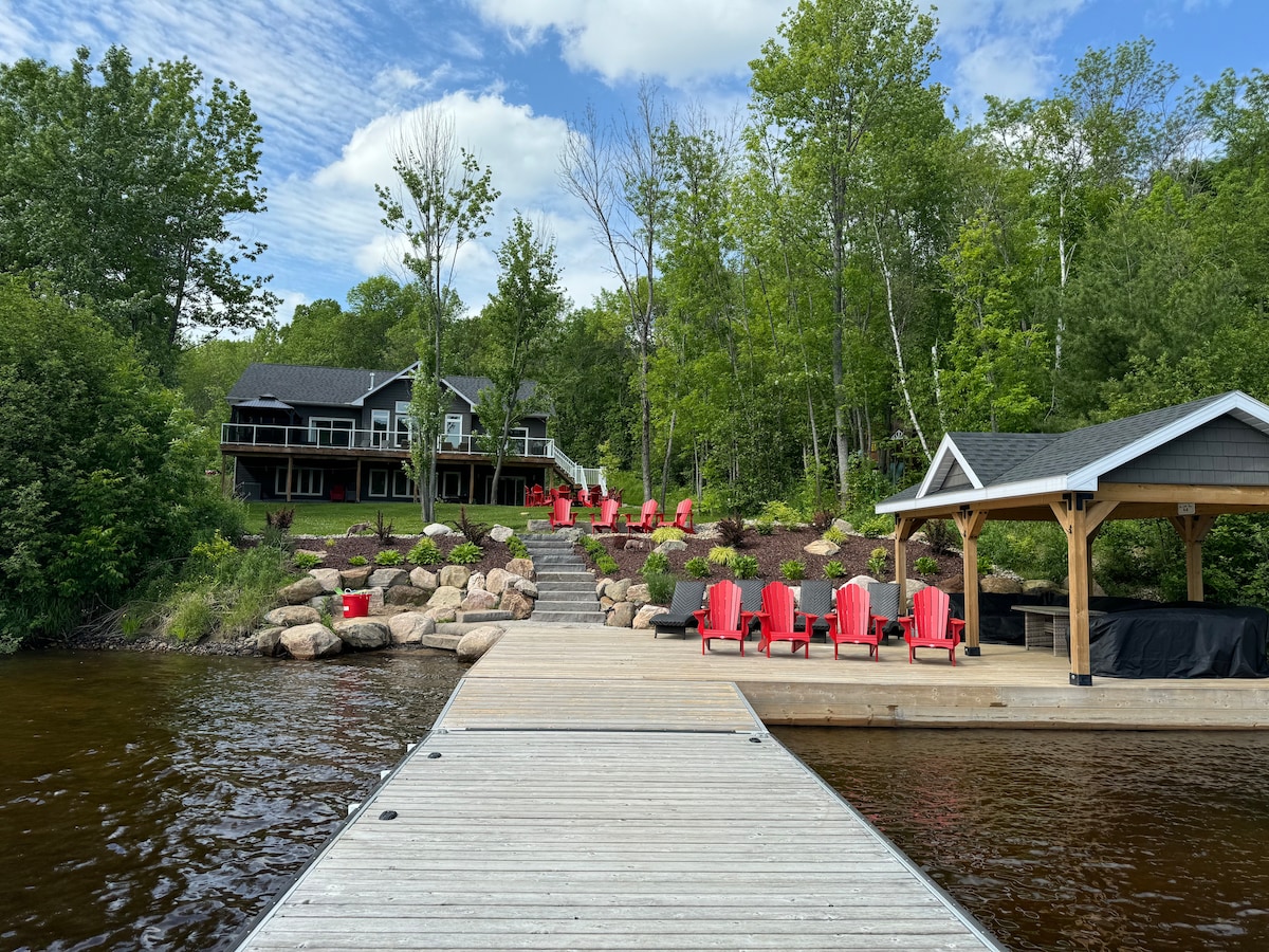 A wooden dock extends into the water, leading to a welcoming gazebo and multiple red Adirondack chairs arranged on the shore. The lush greenery frames the scene, while the two-story cottage is visible in the background, surrounded by natural landscaping.