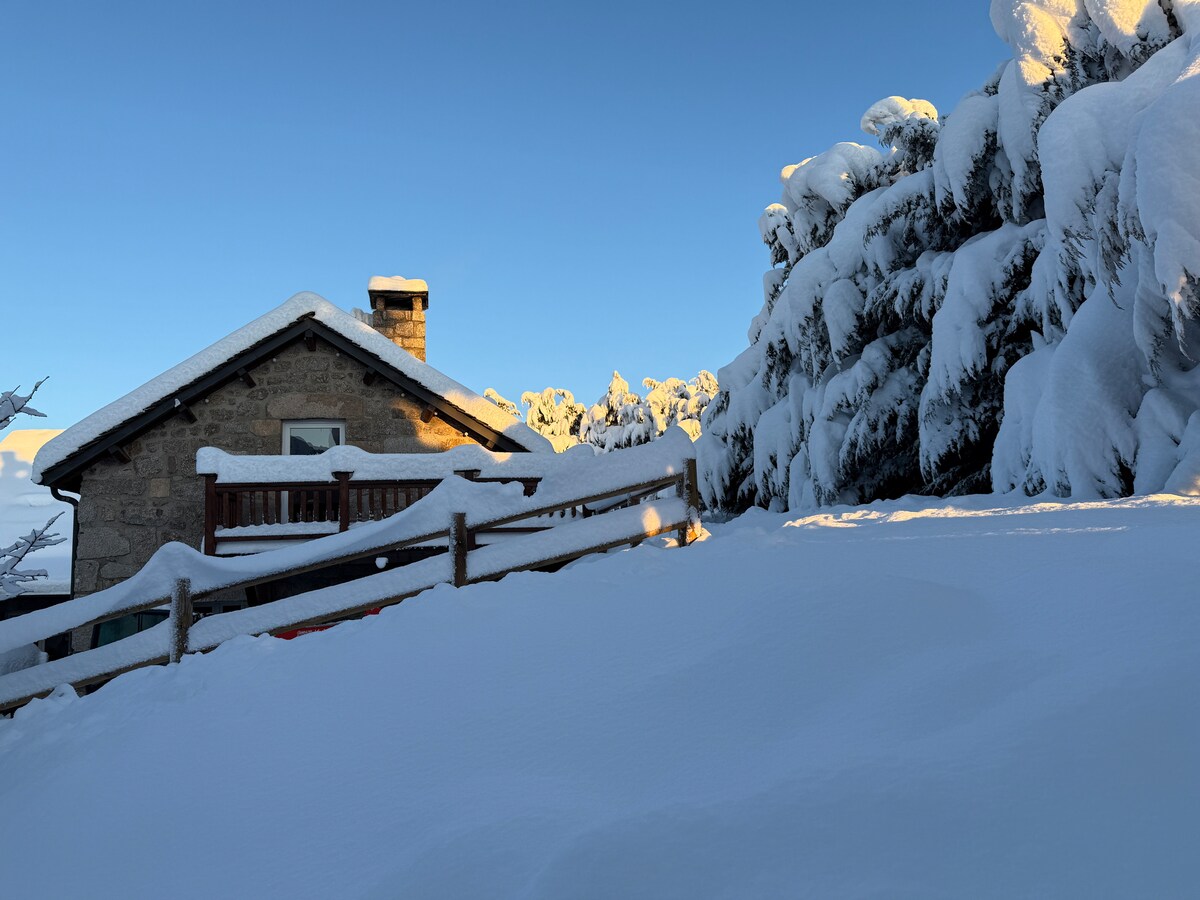 A charming stone exterior is partially covered in snow, contrasting with the bright blue sky. The surrounding landscape is blanketed in fresh snow, with evergreen trees outlined by white. A wooden railing is visible along the pathway, enhancing the serene winter scene.