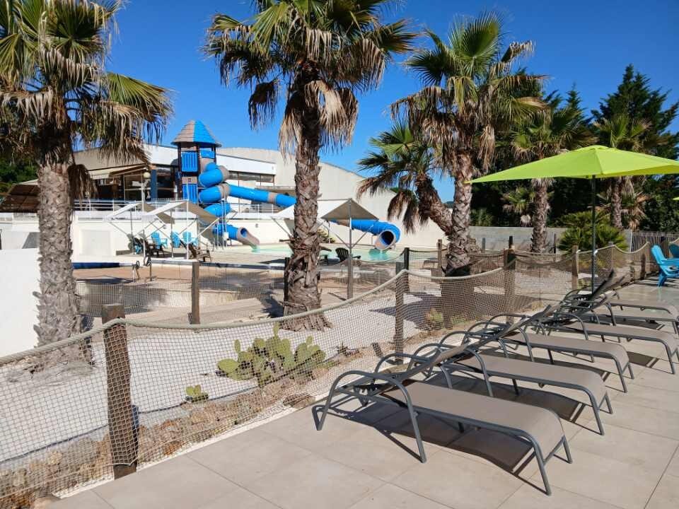 An outdoor pool area features several lounge chairs arranged neatly on a tiled surface. Palm trees provide shade, while a water slide tower is visible in the background. A vibrant green umbrella is set up for additional comfort, enhancing the relaxed atmosphere.