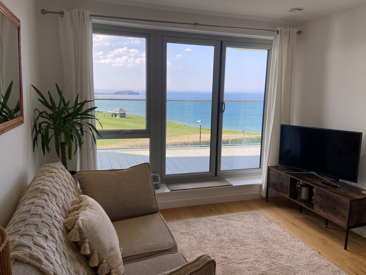A modern lounge area features a comfortable sofa adjacent to a large window, offering expansive views of the coastline and sea. Natural light fills the room, highlighting a neutral color palette. A television is positioned on a low, dark wooden media unit, adding to the contemporary design.