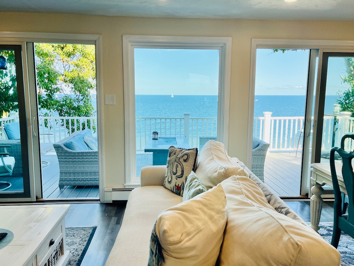 A spacious living area features a comfortable sofa with decorative pillows, facing large glass doors that open to a deck overlooking the ocean. Soft natural light illuminates the room, while the expansive view highlights the serene blue water and sky.