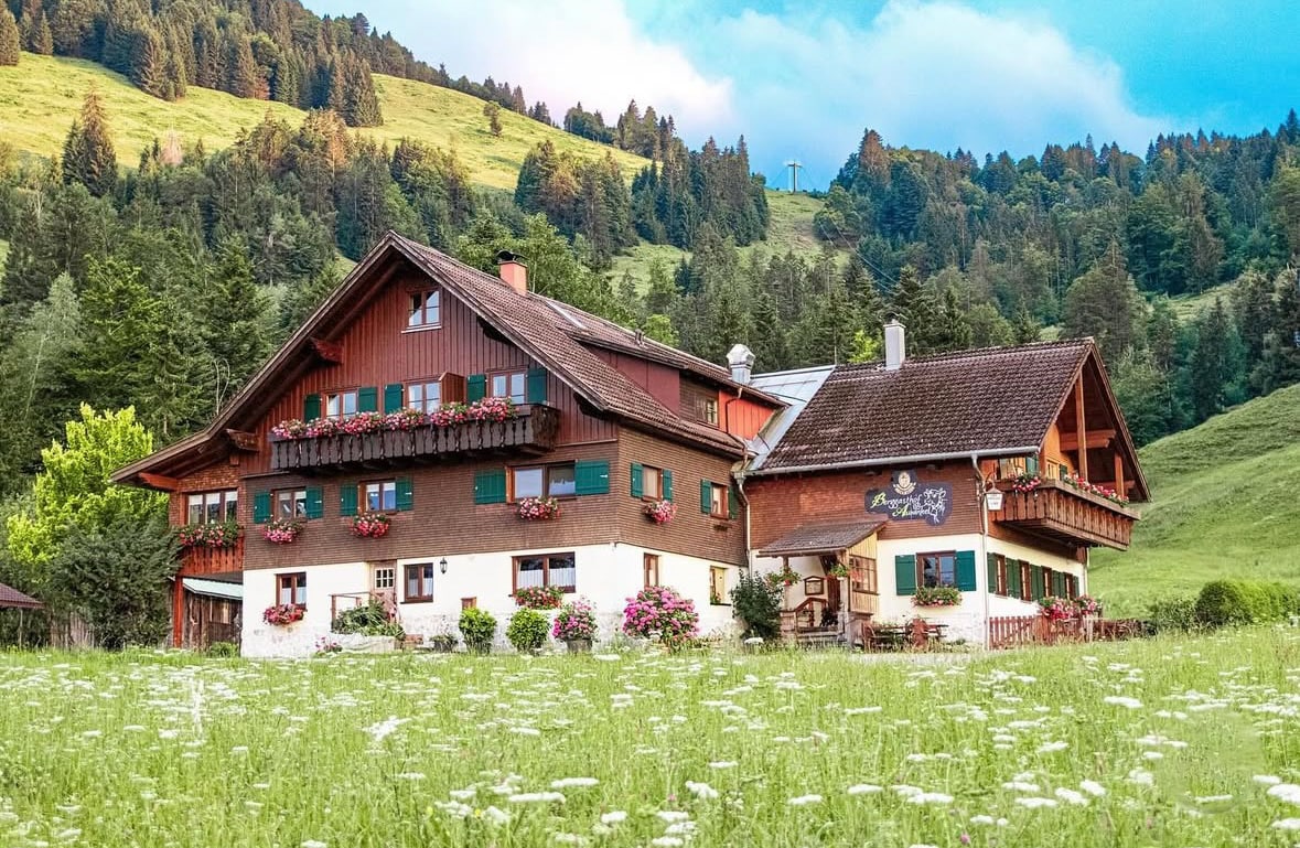 A charming two-story building is nestled amidst lush greenery, featuring wooden balconies adorned with colorful flower boxes. The structure combines traditional wood accents with a warm color palette, set against a backdrop of rolling hills and trees.