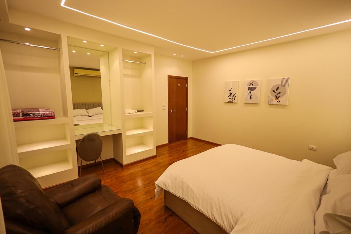 A well-lit bedroom features a spacious bed with white bedding, complemented by a wooden floor. A comfortable chair is positioned beside a sleek desk with a mirror. Decorative wall art adds a subtle touch, while built-in shelving provides additional storage.