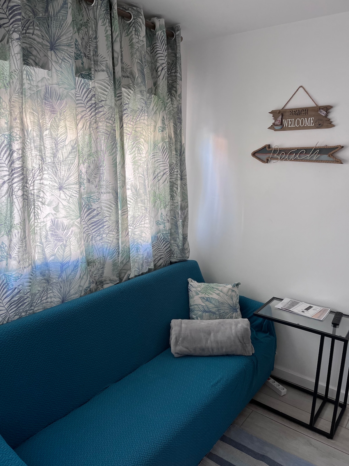 A bright living area features a teal sofa with decorative cushions, accompanied by a sleek side table. Natural light filters through sheer, plant-patterned curtains, enhancing the serene ambiance. A wooden sign with 'Welcome' and 'Beach' adorns the wall, contributing to the coastal theme.