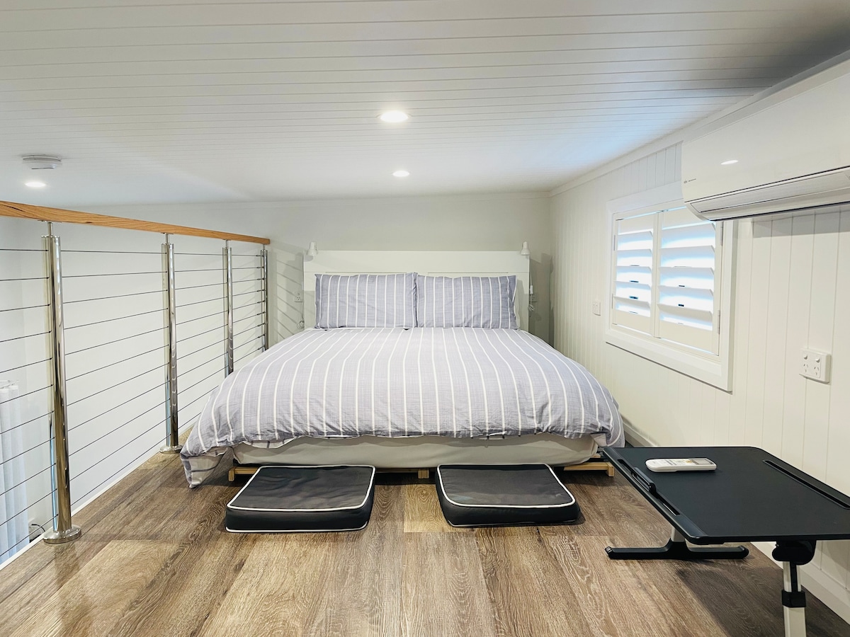 The loft bedroom features a cozy queen bed with a striped duvet, positioned against a wall. A compact desk with an attached tray and two low cushions are situated beside the bed. Natural light enters through a window with shutters.