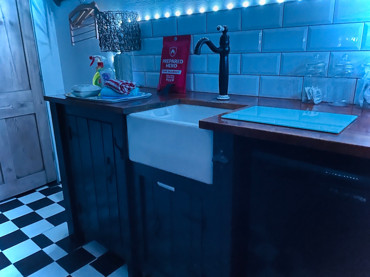 The kitchen area features a hand-built unit with a butler sink, a countertop for meal preparation, and a compact refrigerator. Reflective tiles in soft blue provide a sleek backdrop, while a modern black faucet and ambient lighting enhance visibility.