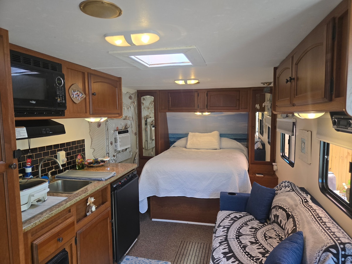 An inviting interior space features a cozy sleeping area with a spacious bed, accented by soft bedding. A well-equipped kitchen is visible, showcasing modern appliances and ample counter space. A small sofa adds comfort, contributing to the functional layout of this compact home.