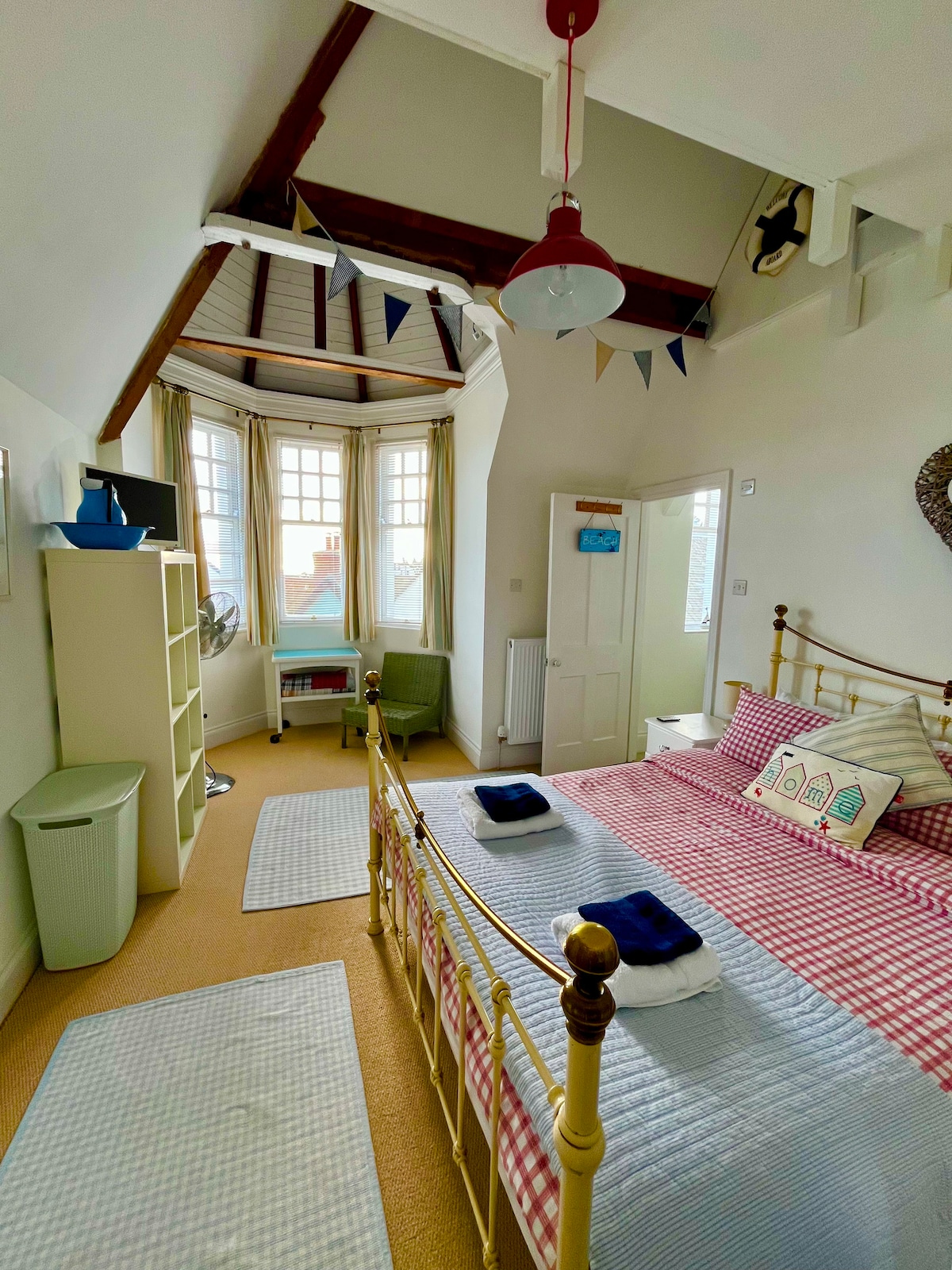 The bedroom features a king-size bed adorned with checkered bedding and two navy towels. Large bay windows allow natural light to fill the space, complemented by decorative bunting along the ceiling. A small desk and chair are positioned near a cozy green armchair.