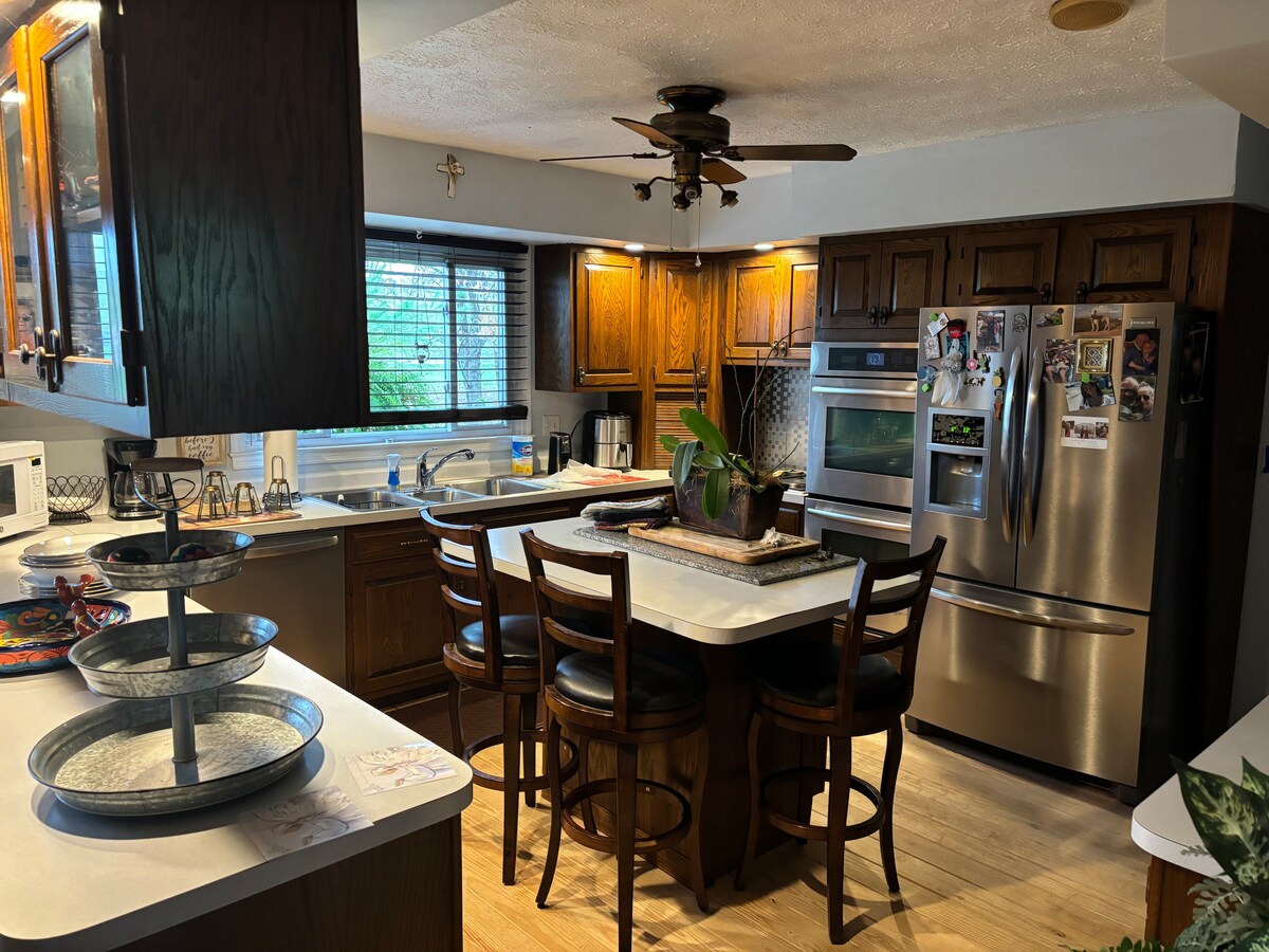 A spacious kitchen is equipped with wooden cabinets and a central island with seating for four. Stainless steel appliances are visible, including an oven and refrigerator. Natural light filters through the window, highlighting decorative elements and an indoor plant among kitchen essentials.