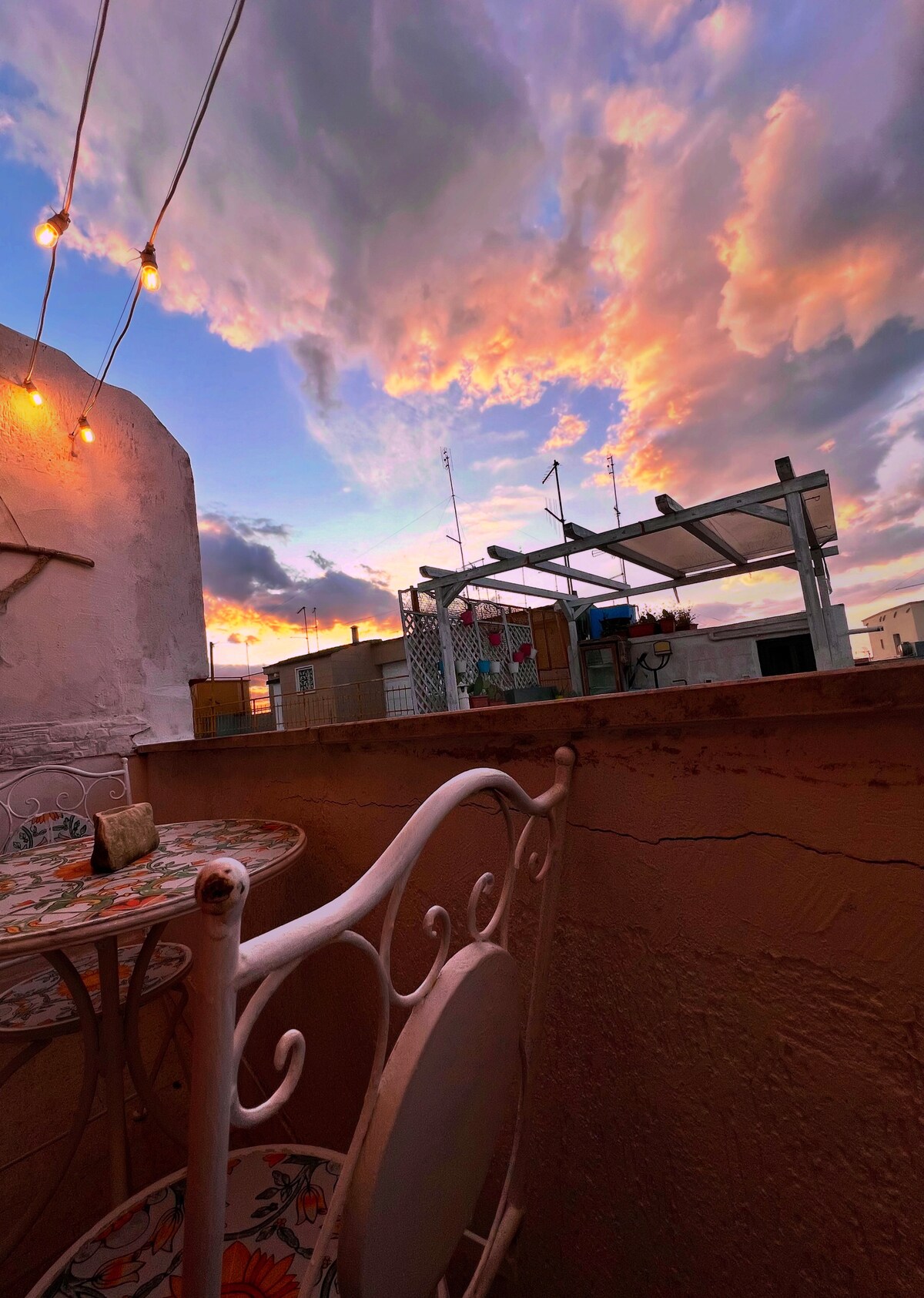 A charming terrace features a small table and iron chairs, illuminated by warming string lights. The sky above displays a breathtaking array of colors as the sun sets, with soft clouds adding depth to the scene. A rooftop structure can be seen in the background.