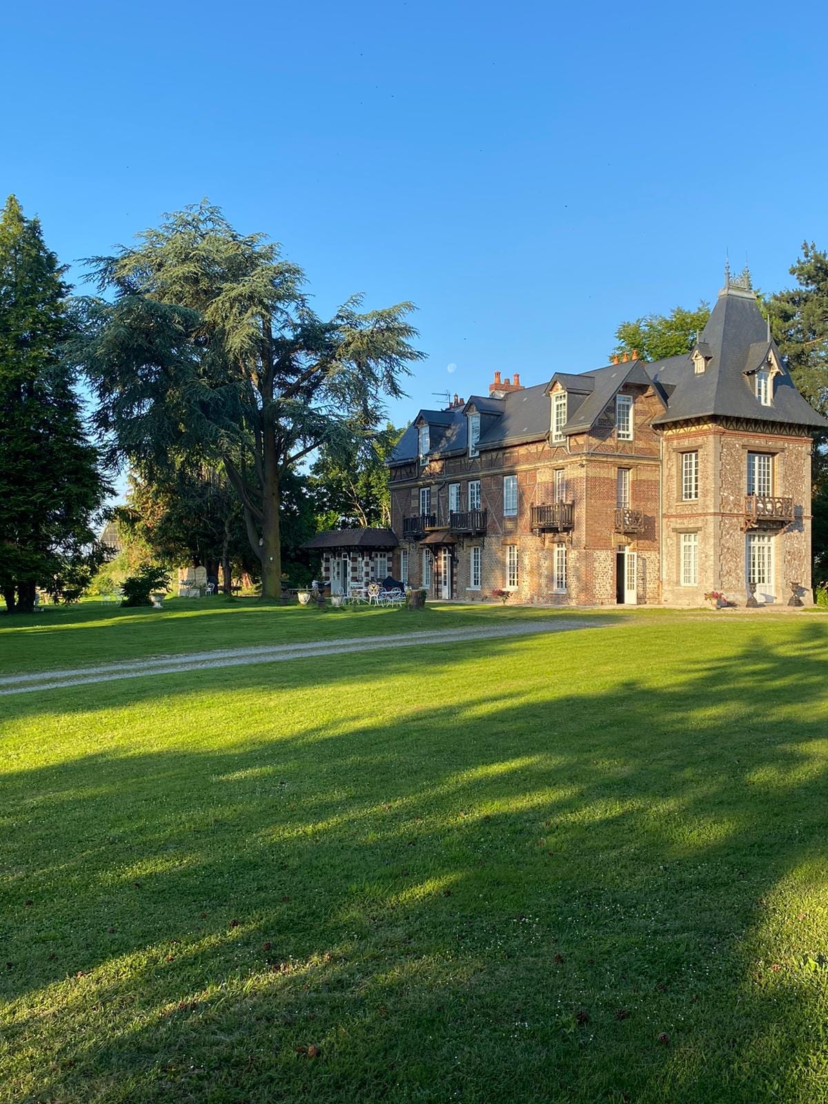 A charming Anglo-Norman manor from 1900 is surrounded by well-maintained parkland and gardens. Its elegant architecture features multiple balconies and peaked roofs. Tall trees cast playful shadows on the expansive green lawn, creating a serene outdoor setting ideal for relaxation.