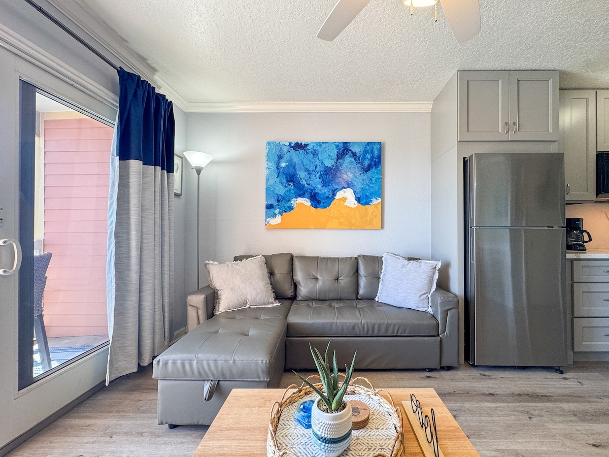The living area features a modern sectional sofa in a neutral tone, complemented by decorative pillows. A vibrant artwork in blue and yellow adorns the wall, contrasting with the light-colored walls. Natural light streams through sliding glass doors, enhanced by simple curtains.