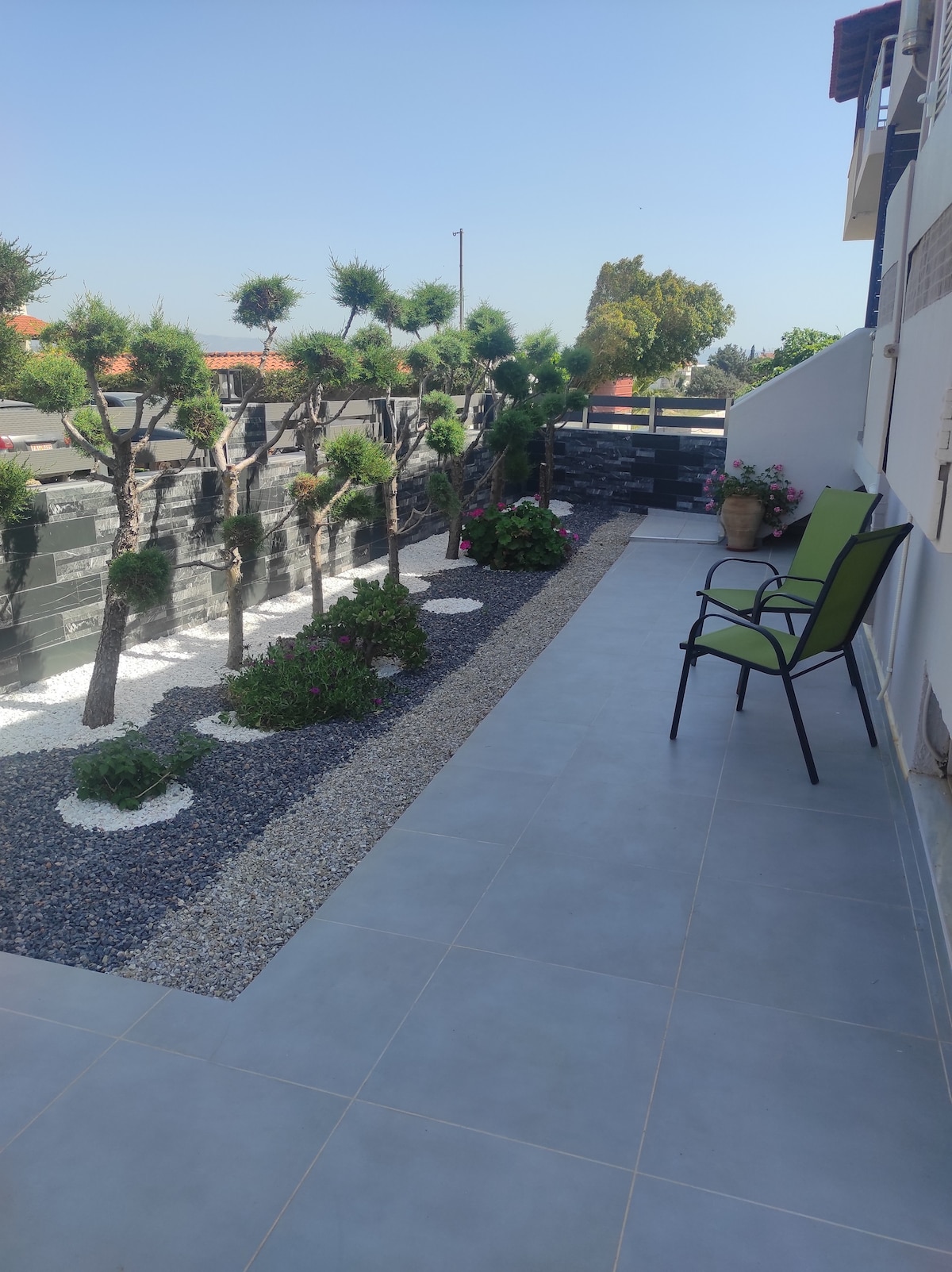 A private outdoor space is shown, featuring a tiled patio with a pair of green chairs positioned next to a manicured garden. The garden includes decorative stones and neatly trimmed shrubs, enhancing the tranquil environment. Clear skies are visible above, contributing to the serene setting.