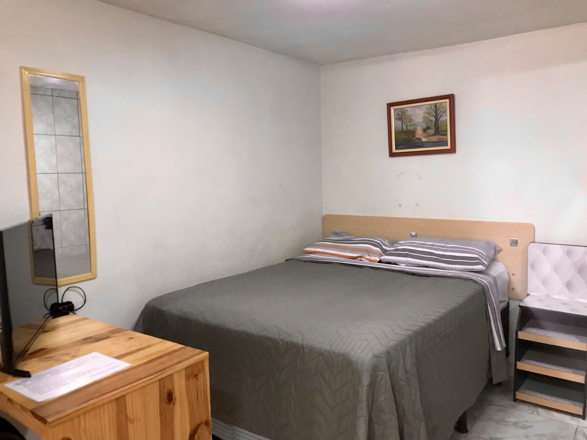 A simple bedroom features a spacious bed with a gray quilt. A small wooden dresser is positioned nearby, while a smart TV is mounted on the wall. Natural light filters in, and a framed landscape painting adds a touch of color.