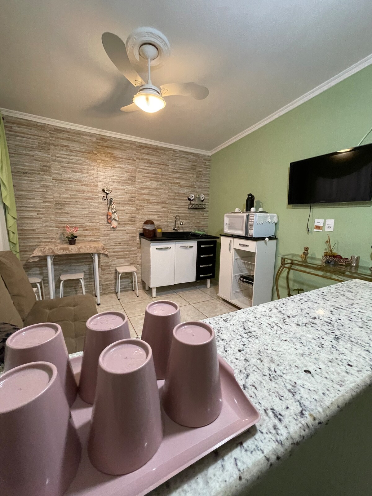 A compact kitchen area features a countertop with a tray holding several pink cups. The kitchen appliances include a microwave and a refrigerator, situated against a textured stone wall. A small table with two chairs is positioned nearby, illuminated by a ceiling fan overhead.