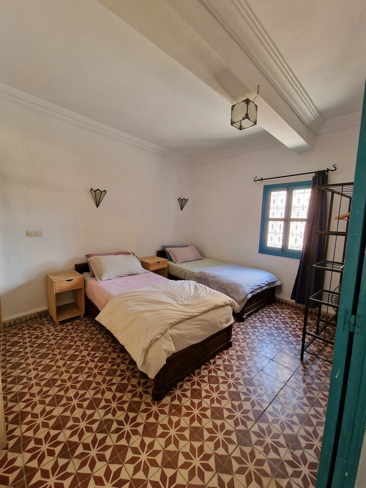 The room features two single beds positioned on either side, with light bedding and wooden frames. Each bed is accompanied by a bedside table. A decorative window with blue frames allows natural light to enter, highlighting the patterned floor tiles.