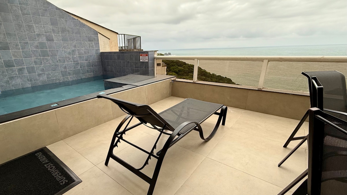 A private balcony area features an inviting heated pool, accompanied by a black lounge chair and a glass table. The backdrop displays an expansive view of the ocean, complemented by a gray stone wall. The space is designed for relaxation.