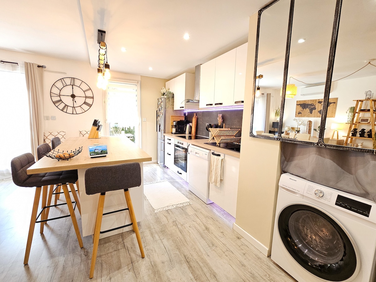 A modern kitchen and dining area are showcased, featuring a wooden table with high-backed chairs. Contemporary appliances, including a washing machine, are visible, alongside decorative elements such as a clock and greenery. Light enters through a window, enhancing the bright and inviting space.
