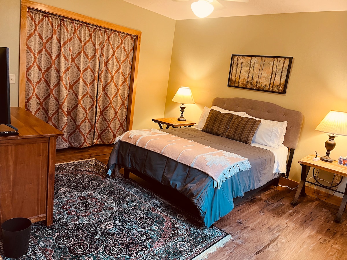 A cozy queen bedroom features a padded headboard with a decorative blanket draped over the bed. A patterned area rug covers the floor, enhancing comfort. Two bedside lamps provide soft lighting, complemented by a window draped with patterned curtains. A small wooden dresser and artwork adorn the walls.