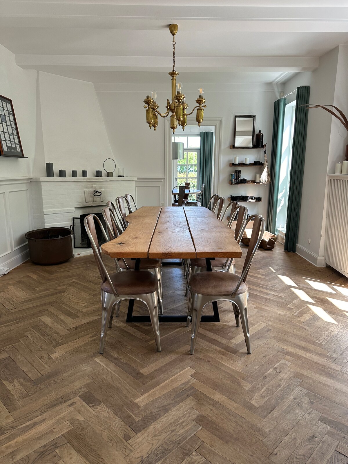 A spacious dining area is highlighted by a large wooden table surrounded by metallic chairs. Natural light fills the room through large windows, illuminating the warm wood flooring. A chandelier hangs overhead, and decorative elements are arranged on the shelves and mantel.