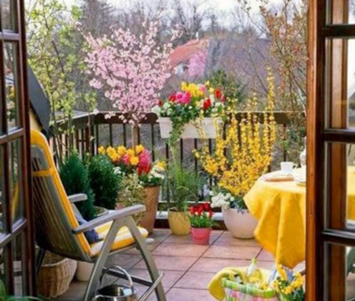 A private terrace is framed by wooden railings and features a sunny arrangement of colorful flowers in various pots. A table covered with a yellow cloth and a comfortable lounge chair invite relaxation amid the greenery and seasonal blooms visible in the background.