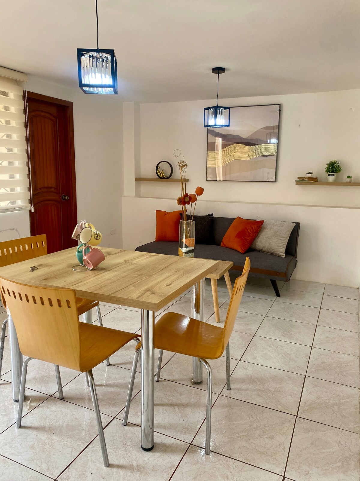 A dining area with a wooden table is surrounded by four light-colored chairs. A small lounge space features a gray sofa adorned with orange cushions. Soft light is provided by two pendant lamps, and a piece of wall art adds a subtle decorative touch.