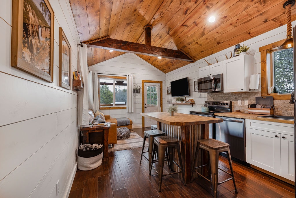 An open-concept kitchen features modern appliances and a wooden island with seating. Natural wood beams accentuate the high ceiling. A cozy living area with a sofa is visible, while a window showcases outdoor greenery, providing ample natural light to the space.