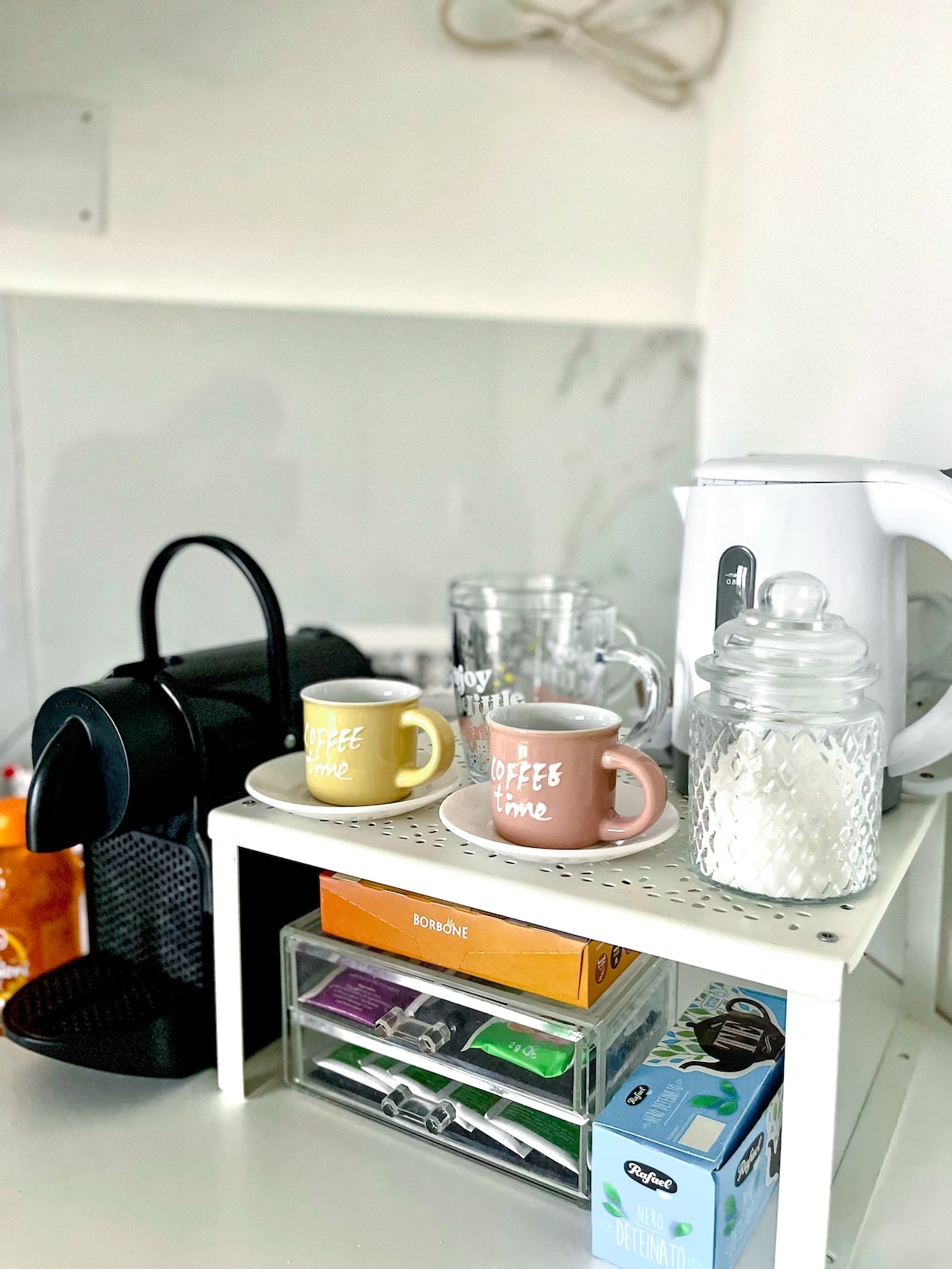 A coffee station features a black coffee machine alongside kettles, mugs, and a glass jar. Two decorative cups are displayed on a white shelf, complemented by various tea bags and sugar packets in an organized drawer below.