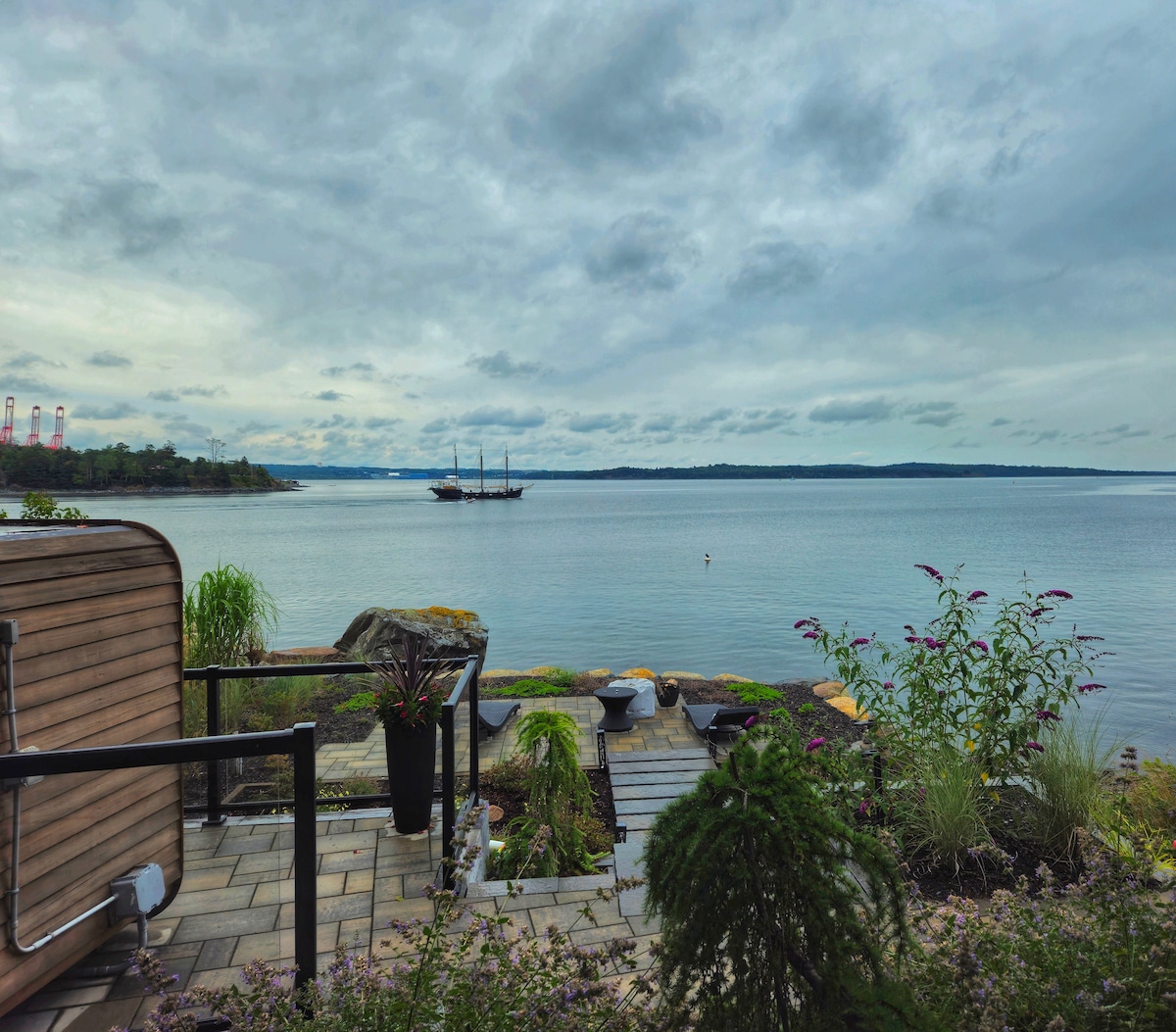 A serene view of the ocean is presented, with gentle waves lapping at the shore. In the foreground, a landscaped area features native plants and stone pathways, leading toward the water. A historic ship is visible in the distance against a cloudy sky.