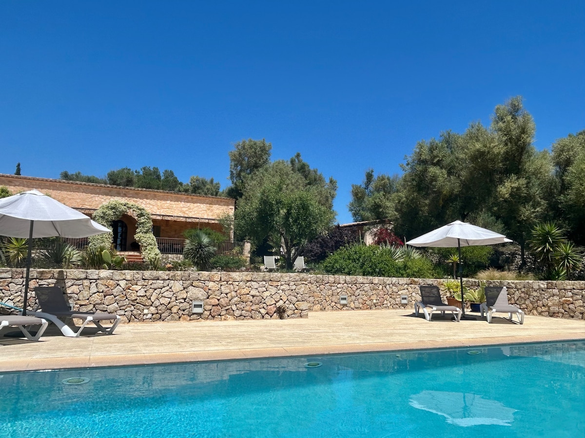 The image depicts a serene pool area featuring a clear blue pool surrounded by sun loungers and white umbrellas. Lush greenery and trees frame the background, showcasing a property built from coral-colored stone. The bright blue sky is visible overhead, enhancing the tranquil setting.
